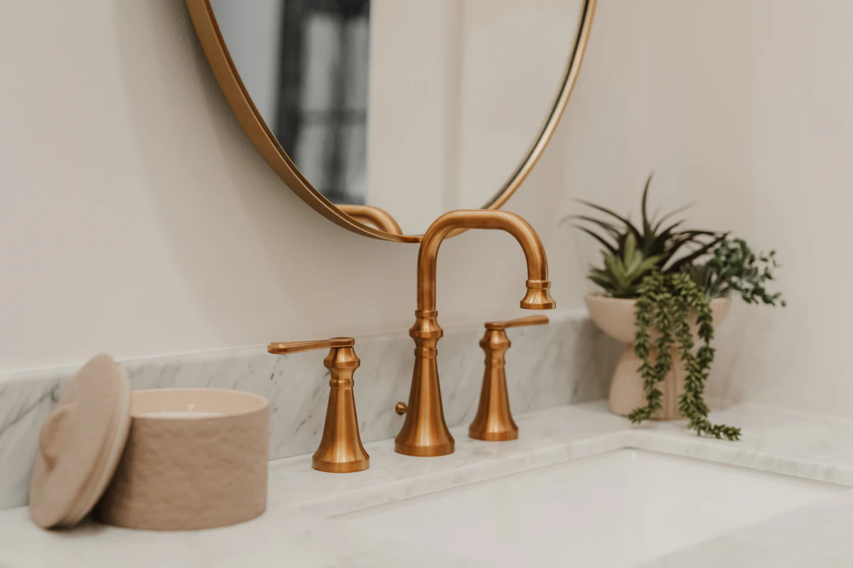 A copper-brass basin tap on a white marble counter beneath a round mirror, with a trailing plant