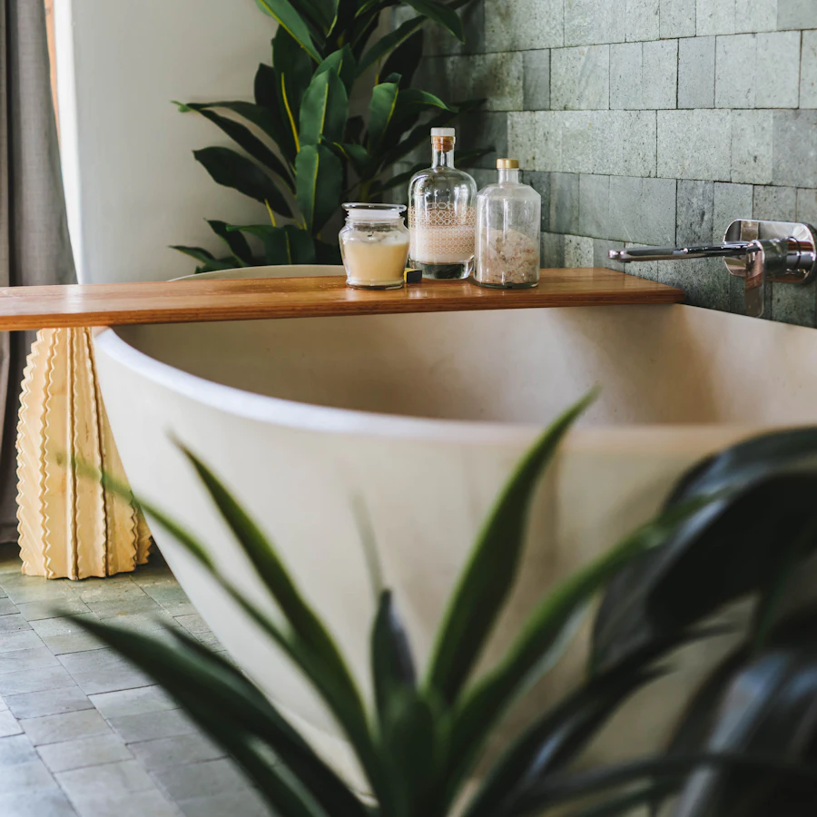 A pale stone bath with a timber shelf holding glass jars, set against green-grey tile among plants