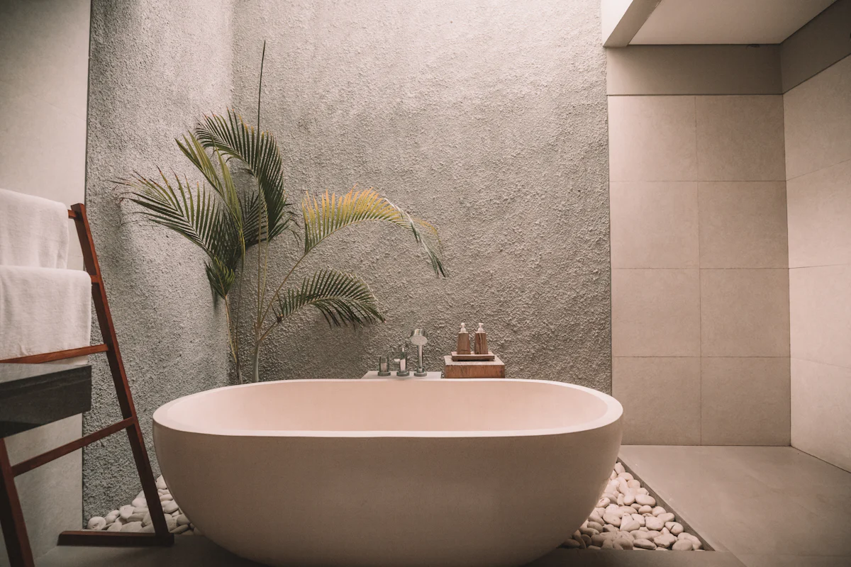 A freestanding stone-coloured bath against a textured plaster wall with a palm and pebble floor