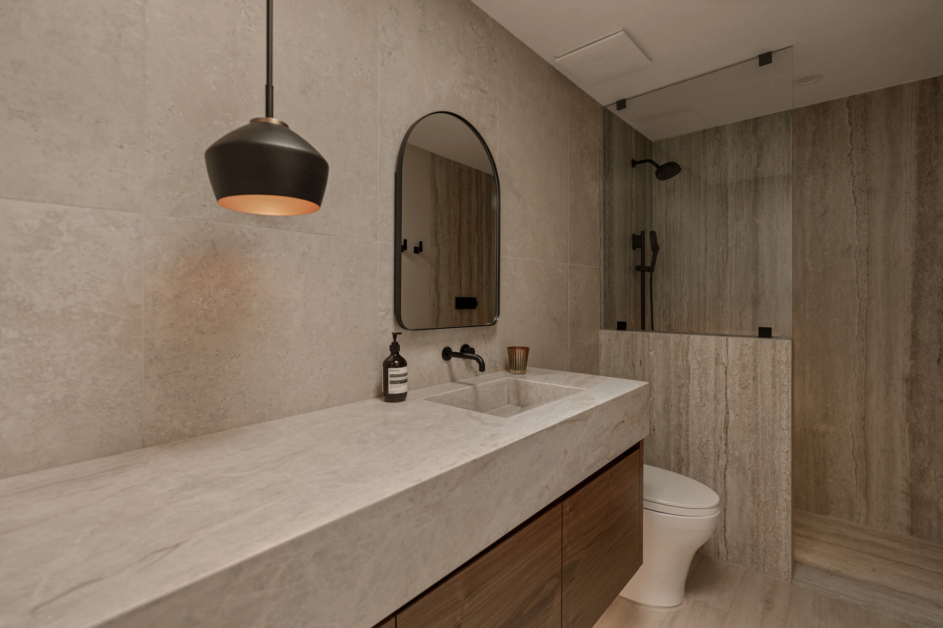 A bathroom in warm grey stone with a long stone vanity, walnut cabinet, arched mirror and a black pendant light