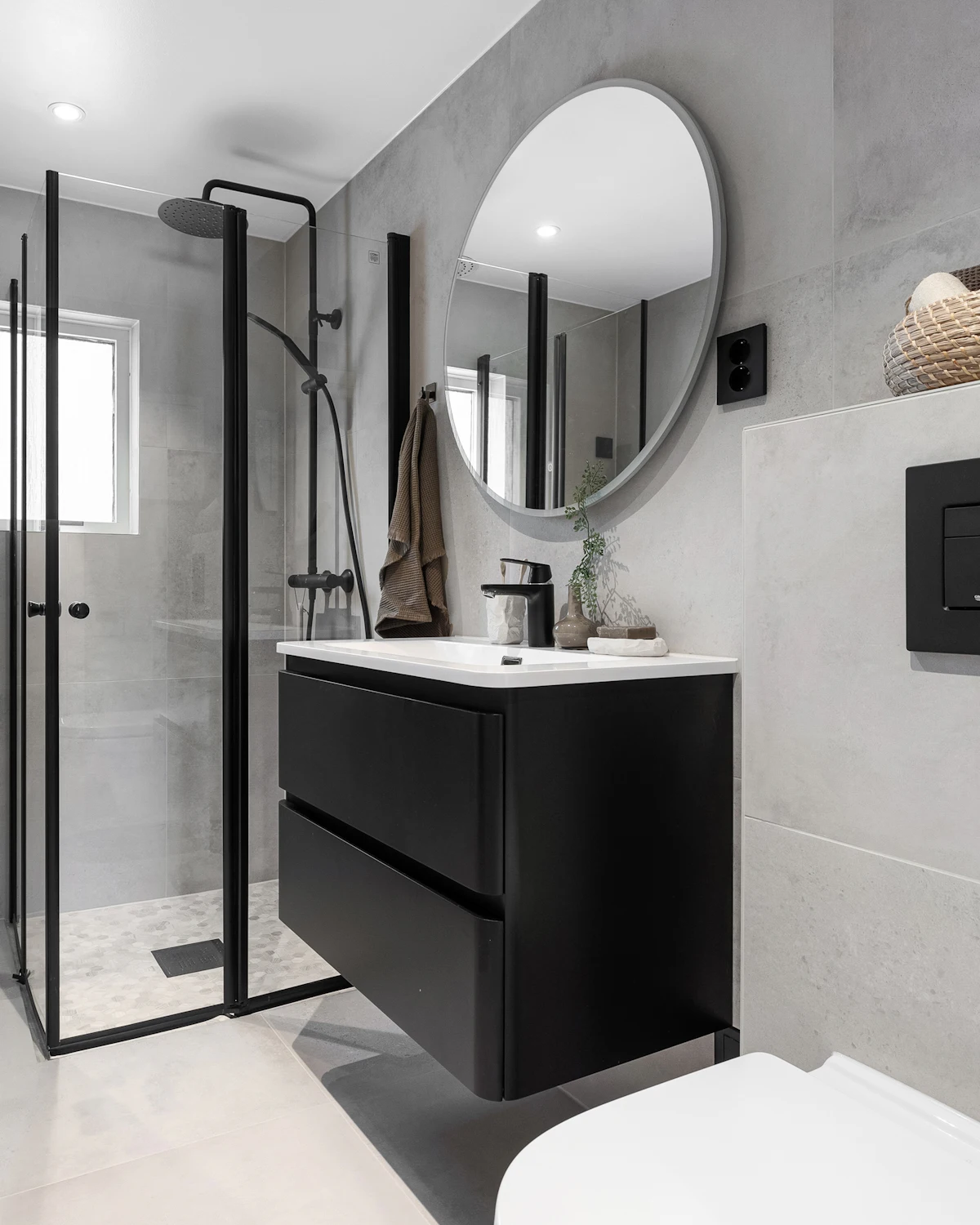 A grey stone-effect bathroom with a matt black wall-hung vanity, round mirror and a black-framed glass shower