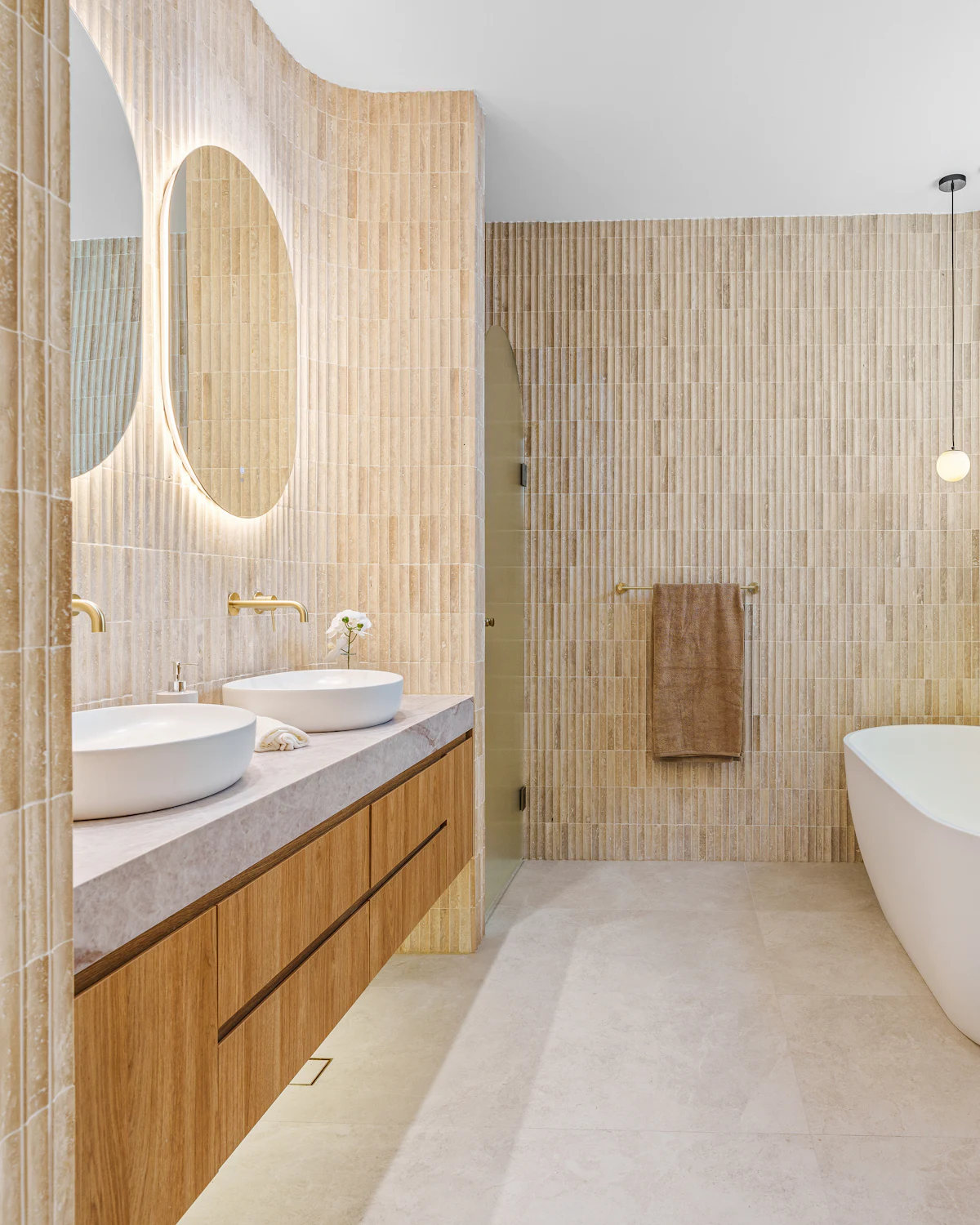 A bathroom with a long oak vanity, two vessel basins, backlit round mirrors, vertical tile and a freestanding bath