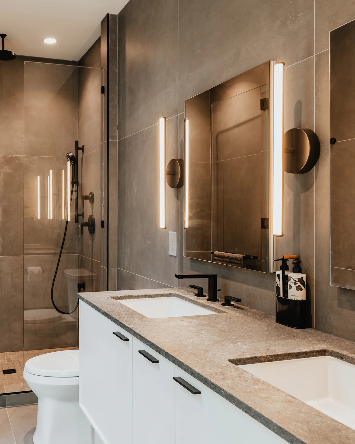 A bathroom with warm brown stone walls, a pale stone counter with two basins, vertical light bars and black fittings